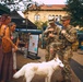 Two U.S. Army soldiers in full camouflage uniforms and tactical gear are interacting with civilians at an outdoor farmers market or street fair. A woman with pink hair and a flowing dress holds a white dog on a leash while conversing with the soldiers. The scene takes place in an urban plaza surrounded by vendor tents and a historic brick building in the background.