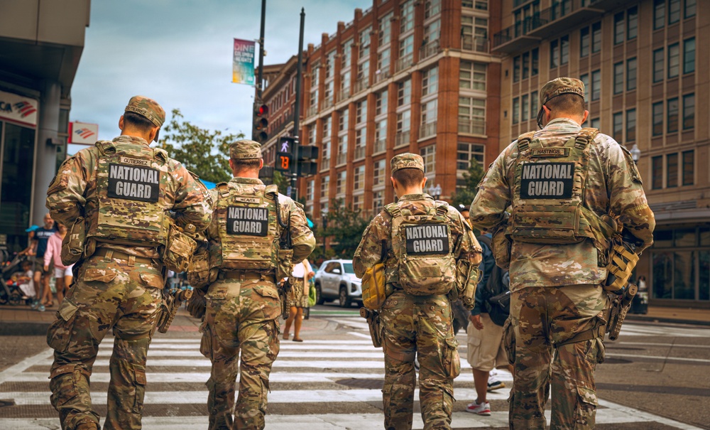 Four National Guard personnel in full camouflage uniforms and tactical vests are walking through a crosswalk in an urban setting. Their backs are visible, showing 'NATIONAL GUARD' patches and name tapes. The scene appears to be in a busy city district with pedestrians, traffic signals, and brick commercial buildings in the background.