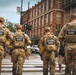 Four National Guard personnel in full camouflage uniforms and tactical vests are walking through a crosswalk in an urban setting. Their backs are visible, showing 'NATIONAL GUARD' patches and name tapes. The scene appears to be in a busy city district with pedestrians, traffic signals, and brick commercial buildings in the background.