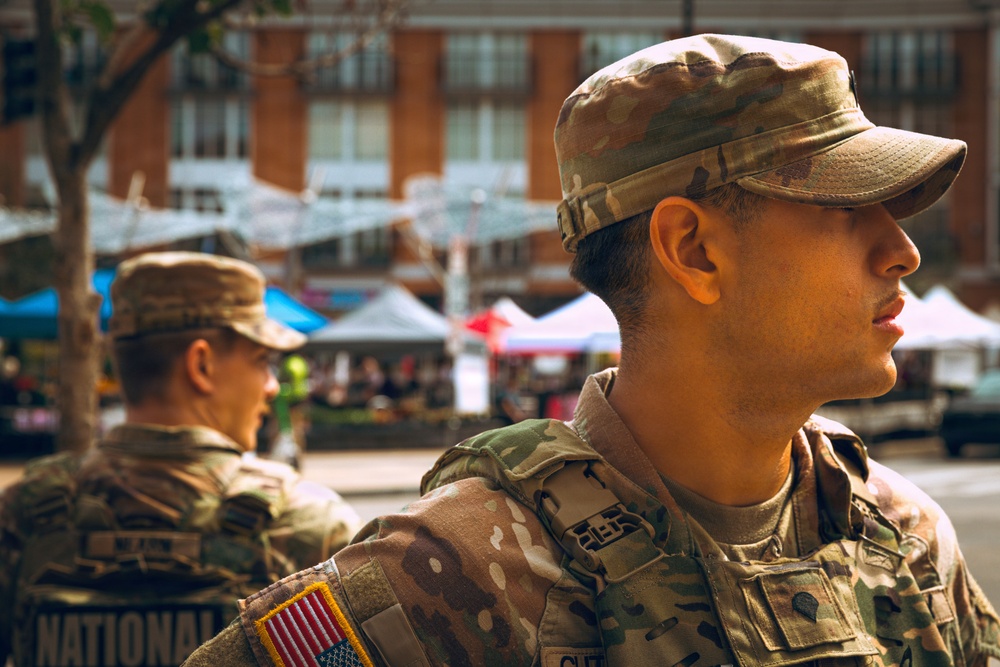 Two soldiers in U.S. Army MultiCam/OCP camouflage uniforms are standing in what appears to be an outdoor public event or market setting. The foreground soldier displays a U.S. flag patch on his left shoulder and a rank/unit insignia on his chest. Partial text on the background soldier's uniform reads 'NATIONAL,' suggesting National Guard affiliation.