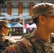 Two soldiers in U.S. Army MultiCam/OCP camouflage uniforms are standing in what appears to be an outdoor public event or market setting. The foreground soldier displays a U.S. flag patch on his left shoulder and a rank/unit insignia on his chest. Partial text on the background soldier's uniform reads 'NATIONAL,' suggesting National Guard affiliation.