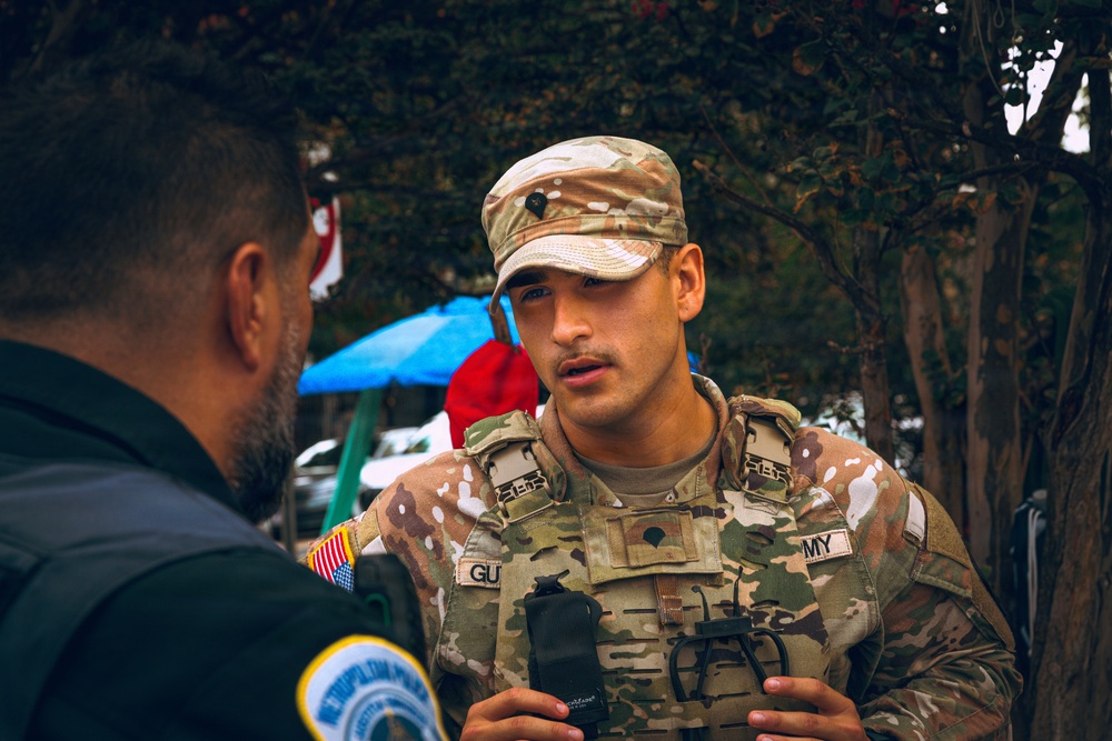 A U.S. Army soldier in MultiCam (OCP) camouflage uniform and tactical vest is engaged in conversation with a law enforcement officer, identifiable by a Metropolitan Police patch on his shoulder. The soldier appears to be at an outdoor event or coordination point, with trees, a blue canopy, and vehicles visible in the background. The interaction suggests inter-agency coordination or community engagement.