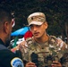A U.S. Army soldier in MultiCam (OCP) camouflage uniform and tactical vest is engaged in conversation with a law enforcement officer, identifiable by a Metropolitan Police patch on his shoulder. The soldier appears to be at an outdoor event or coordination point, with trees, a blue canopy, and vehicles visible in the background. The interaction suggests inter-agency coordination or community engagement.