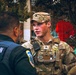 A U.S. Army soldier wearing OCP (Operational Camouflage Pattern) uniform with a Sergeant rank insignia and body armor is engaged in conversation with a Metropolitan Police officer. The interaction appears to be a coordination or briefing between military and law enforcement personnel in an outdoor urban setting. A third individual in military uniform is partially visible on the right side of the frame.