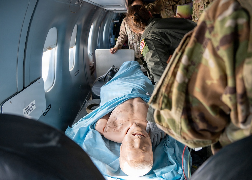 Military medical personnel in OCP (Operational Camouflage Pattern) uniforms are conducting a medical training exercise aboard an aircraft. A realistic medical training mannequin is positioned on a litter/stretcher, covered with a blue drape, simulating a casualty scenario. The setting appears to be an aeromedical evacuation training exercise based on the aircraft interior and military uniforms with U.S. flag patches.