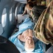 Military medical personnel in OCP (Operational Camouflage Pattern) uniforms are conducting a medical training exercise aboard an aircraft. A realistic medical training mannequin is positioned on a litter/stretcher, covered with a blue drape, simulating a casualty scenario. The setting appears to be an aeromedical evacuation training exercise based on the aircraft interior and military uniforms with U.S. flag patches.