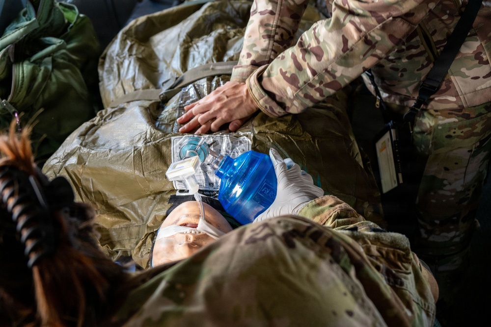 Military medical personnel in MultiCam/OCP camouflage uniforms are performing emergency medical care on a casualty. One provider is administering ventilation using a bag-valve mask (BVM) while another is performing chest compressions or assessment. This appears to be a tactical combat casualty care (TCCC) scenario, either a training exercise or real medical response.
