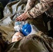Military medical personnel in MultiCam/OCP camouflage uniforms are performing emergency medical care on a casualty. One provider is administering ventilation using a bag-valve mask (BVM) while another is performing chest compressions or assessment. This appears to be a tactical combat casualty care (TCCC) scenario, either a training exercise or real medical response.