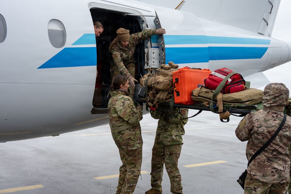 Military personnel in camouflage uniforms are loading medical and tactical equipment onto a white aircraft with blue stripe markings on a wet tarmac. The team appears to be conducting an aeromedical evacuation or rapid deployment operation, transferring a litter (stretcher) loaded with orange Pelican cases, tactical bags, and red medical kits into the aircraft door. The overcast, rainy conditions suggest an urgent or operational deployment scenario.