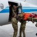 Military personnel in camouflage uniforms are loading medical and tactical equipment onto a white aircraft with blue stripe markings on a wet tarmac. The team appears to be conducting an aeromedical evacuation or rapid deployment operation, transferring a litter (stretcher) loaded with orange Pelican cases, tactical bags, and red medical kits into the aircraft door. The overcast, rainy conditions suggest an urgent or operational deployment scenario.