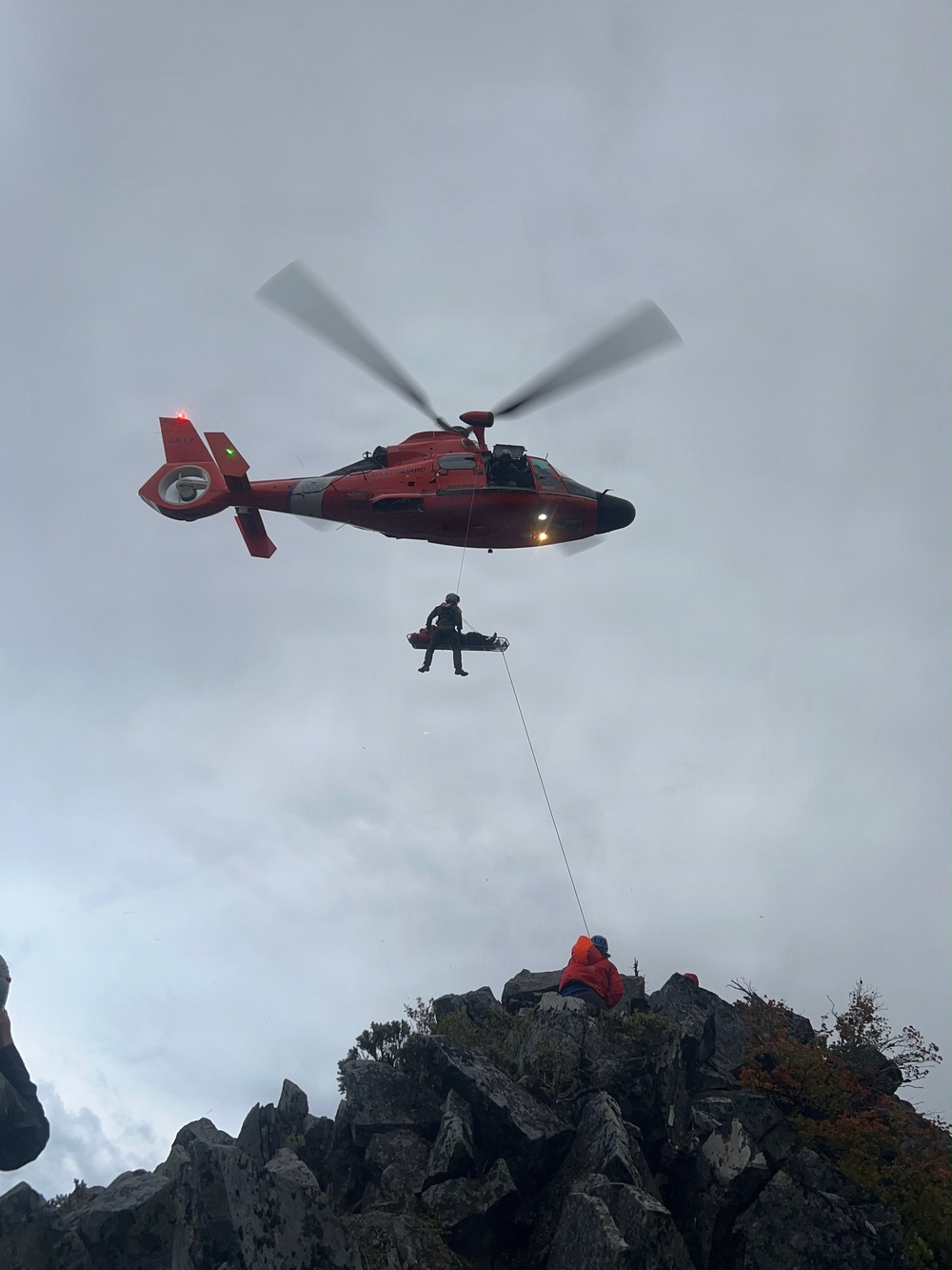 A U.S. Coast Guard helicopter (orange/red MH-65 Dolphin) is performing a hoist rescue operation over a rocky mountain ridge. A rescue swimmer is being lifted via hoist cable while accompanying a patient on a litter/stretcher. At least one person in an orange jacket remains on the rocky outcrop below, likely a ground rescue team member managing a tag line.