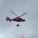 A U.S. Coast Guard helicopter (orange/red MH-65 Dolphin) is performing a hoist rescue operation over a rocky mountain ridge. A rescue swimmer is being lifted via hoist cable while accompanying a patient on a litter/stretcher. At least one person in an orange jacket remains on the rocky outcrop below, likely a ground rescue team member managing a tag line.