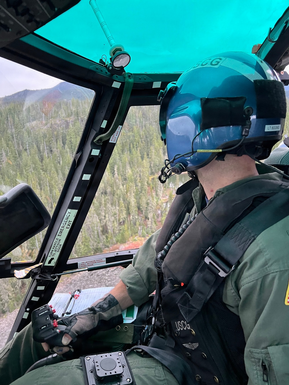 A United States Coast Guard (USCG) pilot is seated in the cockpit of a helicopter, flying over a densely forested mountainous terrain. The pilot is wearing a teal/blue flight helmet marked 'USCG' and a label reading 'LT BOOKS', along with a green flight suit and tactical vest also marked 'USCG'. The helicopter appears to be on a search and rescue or patrol mission over what appears to be a Pacific Northwest forest landscape.