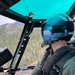 A United States Coast Guard (USCG) pilot is seated in the cockpit of a helicopter, flying over a densely forested mountainous terrain. The pilot is wearing a teal/blue flight helmet marked 'USCG' and a label reading 'LT BOOKS', along with a green flight suit and tactical vest also marked 'USCG'. The helicopter appears to be on a search and rescue or patrol mission over what appears to be a Pacific Northwest forest landscape.