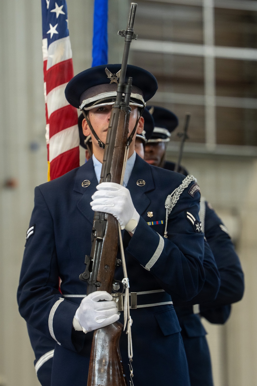 United States Air Force honor guard members stand at attention during a formal military ceremony. The foreground airman holds an M1 Garand rifle in a ceremonial position, wearing the distinctive dark blue dress uniform with white gloves and service cap. An American flag and a blue Air Force flag are visible in the background.