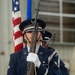 United States Air Force honor guard members stand at attention during a formal military ceremony. The foreground airman holds an M1 Garand rifle in a ceremonial position, wearing the distinctive dark blue dress uniform with white gloves and service cap. An American flag and a blue Air Force flag are visible in the background.
