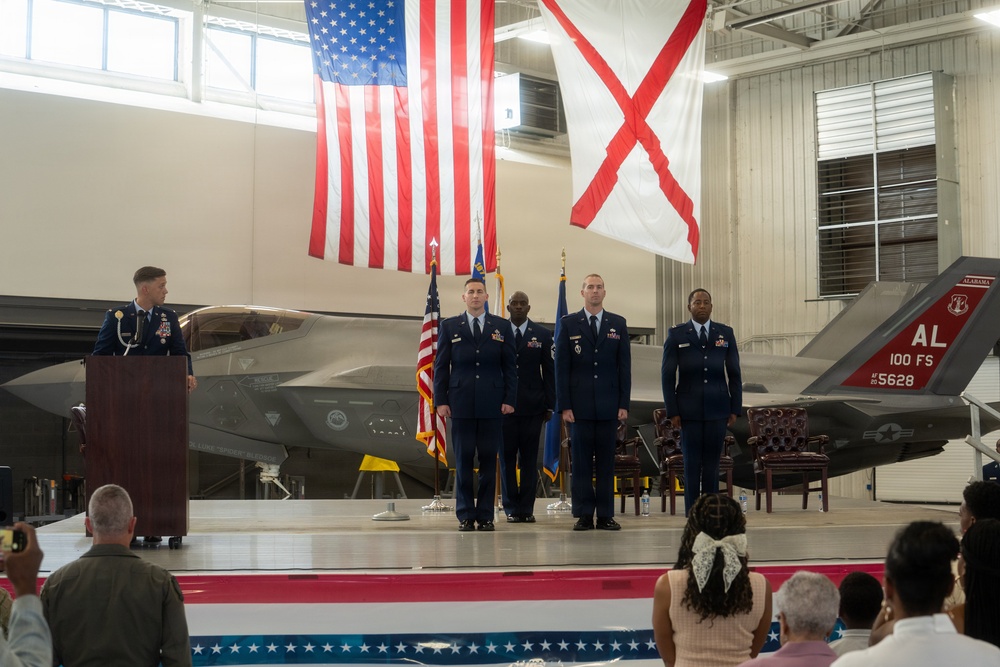 A formal military ceremony is taking place inside an aircraft hangar, with uniformed Air Force personnel standing at attention on a stage. The backdrop features two F-35 fighter jets, a large American flag, and the Alabama state flag. A senior officer is speaking at a podium while three service members stand in formation, suggesting a change of command or promotion ceremony.