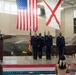 A formal military ceremony is taking place inside an aircraft hangar, with uniformed Air Force personnel standing at attention on a stage. The backdrop features two F-35 fighter jets, a large American flag, and the Alabama state flag. A senior officer is speaking at a podium while three service members stand in formation, suggesting a change of command or promotion ceremony.