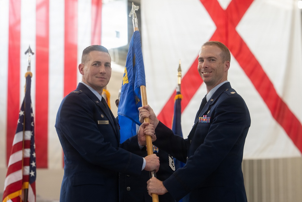 Two U.S. Air Force officers are participating in a formal change of command ceremony, passing a unit guidon (flag) between them. The ceremony is taking place in what appears to be a hangar or large indoor venue decorated with the U.S. flag and what appears to be the Alabama state flag. Both officers are in dress blue uniforms, with the officer on the right displaying multiple service ribbons.