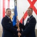 Two U.S. Air Force officers are participating in a formal change of command ceremony, passing a unit guidon (flag) between them. The ceremony is taking place in what appears to be a hangar or large indoor venue decorated with the U.S. flag and what appears to be the Alabama state flag. Both officers are in dress blue uniforms, with the officer on the right displaying multiple service ribbons.