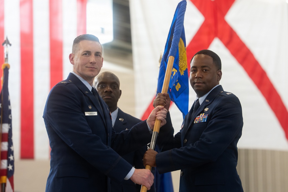 Two U.S. Air Force officers are participating in a formal change of command ceremony, jointly holding a unit guidon (flag on a wooden staff). A third uniformed service member stands in the background. The ceremony appears to be taking place inside a hangar or large military facility decorated with the U.S. flag and what appears to be the Alabama state flag.