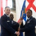 Two U.S. Air Force officers are participating in a formal change of command ceremony, jointly holding a unit guidon (flag on a wooden staff). A third uniformed service member stands in the background. The ceremony appears to be taking place inside a hangar or large military facility decorated with the U.S. flag and what appears to be the Alabama state flag.