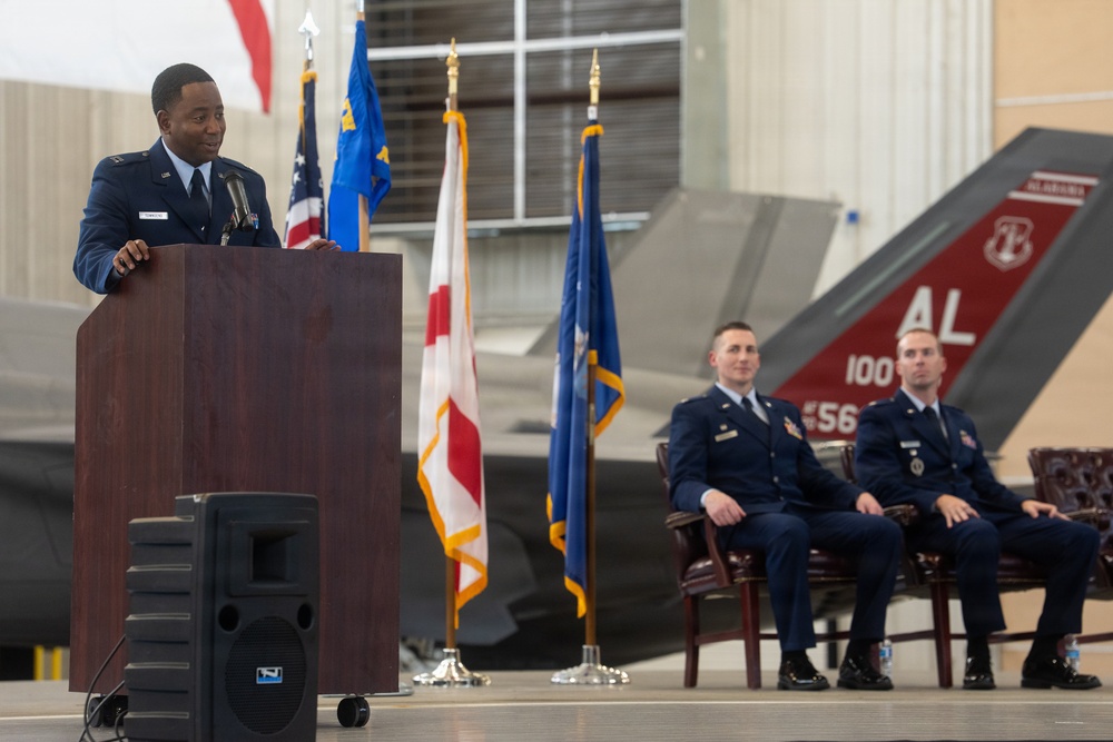 A U.S. Air Force officer is delivering a speech at a podium during a formal military ceremony held inside an aircraft hangar. Two other uniformed Air Force officers are seated in chairs behind him, with multiple flags displayed including the U.S. flag, Air Force flag, and what appears to be the Alabama state flag. A military aircraft, likely an F-35, is visible in the background with 'AL' markings on its tail.