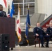 A U.S. Air Force officer is delivering a speech at a podium during a formal military ceremony held inside an aircraft hangar. Two other uniformed Air Force officers are seated in chairs behind him, with multiple flags displayed including the U.S. flag, Air Force flag, and what appears to be the Alabama state flag. A military aircraft, likely an F-35, is visible in the background with 'AL' markings on its tail.