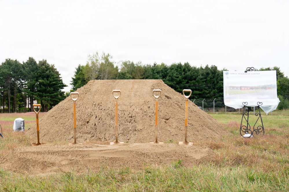 This image depicts a groundbreaking ceremony setup at an outdoor construction site. Five ceremonial gold-painted shovels are planted in a mound of dirt, a common tradition marking the start of a new construction project. A display board on an ornamental easel to the right reads 'Family Readiness Center,' indicating this is a military or community facility groundbreaking.
