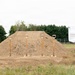 This image depicts a groundbreaking ceremony setup at an outdoor construction site. Five ceremonial gold-painted shovels are planted in a mound of dirt, a common tradition marking the start of a new construction project. A display board on an ornamental easel to the right reads 'Family Readiness Center,' indicating this is a military or community facility groundbreaking.