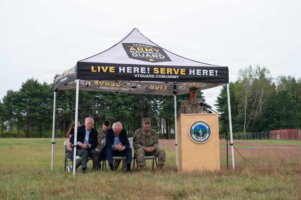 An outdoor ceremony is taking place under a Vermont Army National Guard branded canopy tent on a grassy field. Several individuals, including civilians in suits and military personnel in camouflage uniforms, are seated while a uniformed soldier speaks at a podium bearing an official seal. The event appears to be a formal military or civic function associated with the Vermont Army National Guard.