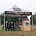 An outdoor ceremony is taking place under a Vermont Army National Guard branded canopy tent on a grassy field. Several individuals, including civilians in suits and military personnel in camouflage uniforms, are seated while a uniformed soldier speaks at a podium bearing an official seal. The event appears to be a formal military or civic function associated with the Vermont Army National Guard.