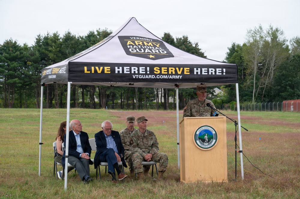 An outdoor ceremony or event is being held under a Vermont Army National Guard branded canopy tent in a grassy field. A uniformed military officer is speaking at a podium bearing the Vermont National Guard seal, while several seated attendees — including two civilians in suits and two military personnel in camouflage uniforms — listen. The event appears to be an official military or civic function.