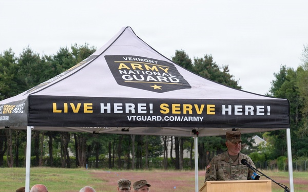 An outdoor ceremony or event is being held under a Vermont Army National Guard branded canopy tent in a grassy field. A uniformed military officer is speaking at a podium bearing the Vermont National Guard seal, while several seated attendees — including two civilians in suits and two military personnel in camouflage uniforms — listen. The event appears to be an official military or civic function.