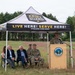 An outdoor ceremony or event is being held under a Vermont Army National Guard branded canopy tent in a grassy field. A uniformed military officer is speaking at a podium bearing the Vermont National Guard seal, while several seated attendees — including two civilians in suits and two military personnel in camouflage uniforms — listen. The event appears to be an official military or civic function.
