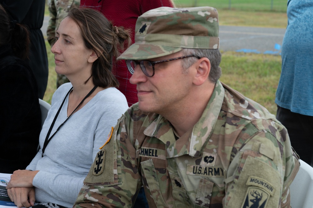 A U.S. Army officer wearing OCP (Operational Camouflage Pattern) uniform is seated alongside a civilian woman at what appears to be an outdoor military event or briefing. The soldier displays insignia identifying him as a Lieutenant Colonel assigned to the 10th Mountain Division. Both individuals appear to be attentively observing something off-camera.