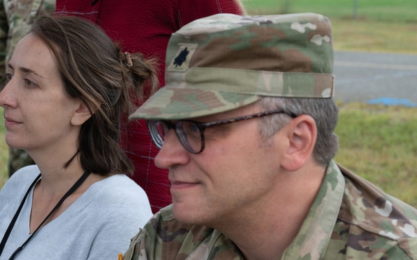 A U.S. Army officer wearing OCP (Operational Camouflage Pattern) uniform is seated alongside a civilian woman at what appears to be an outdoor military event or briefing. The soldier displays insignia identifying him as a Lieutenant Colonel assigned to the 10th Mountain Division. Both individuals appear to be attentively observing something off-camera.