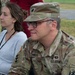A U.S. Army officer wearing OCP (Operational Camouflage Pattern) uniform is seated alongside a civilian woman at what appears to be an outdoor military event or briefing. The soldier displays insignia identifying him as a Lieutenant Colonel assigned to the 10th Mountain Division. Both individuals appear to be attentively observing something off-camera.