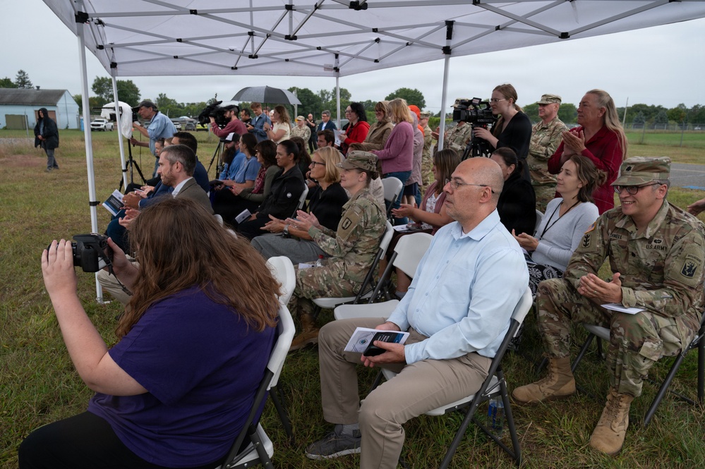 An outdoor ceremony or event is taking place under a white canopy tent on a grassy field during overcast/rainy weather. The audience consists of a mix of military personnel in camouflage uniforms and civilians seated in white folding chairs. Media personnel with cameras are present, suggesting this is an official or newsworthy event.