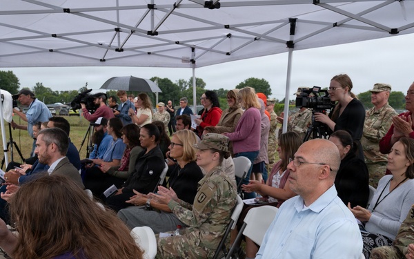 An outdoor ceremony or event is taking place under a white canopy tent on a grassy field during overcast/rainy weather. The audience consists of a mix of military personnel in camouflage uniforms and civilians seated in white folding chairs. Media personnel with cameras are present, suggesting this is an official or newsworthy event.
