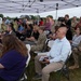 An outdoor ceremony or event is taking place under a white canopy tent on a grassy field during overcast/rainy weather. The audience consists of a mix of military personnel in camouflage uniforms and civilians seated in white folding chairs. Media personnel with cameras are present, suggesting this is an official or newsworthy event.