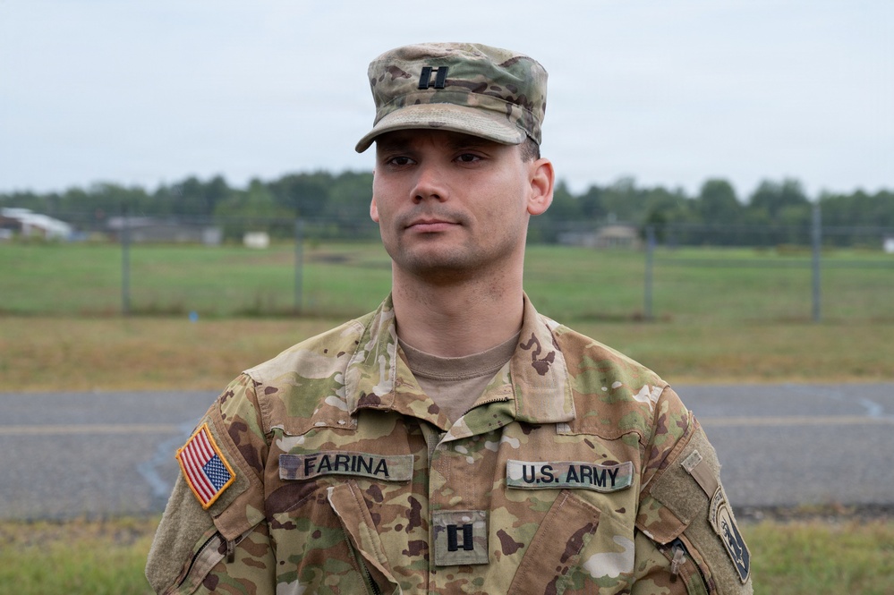 A U.S. Army Captain named Farina is photographed in a formal portrait-style image while wearing Operational Camouflage Pattern (OCP) uniform. He is standing outdoors on what appears to be a military installation, with an open field and tree line visible in the background. His rank insignia (Captain's double bars) is visible on both his patrol cap and chest.