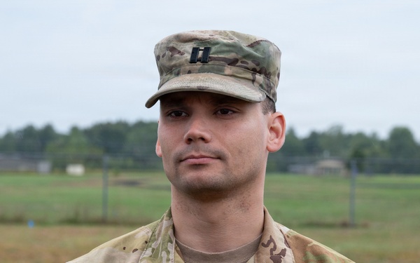 A U.S. Army Captain named Farina is photographed in a formal portrait-style image while wearing Operational Camouflage Pattern (OCP) uniform. He is standing outdoors on what appears to be a military installation, with an open field and tree line visible in the background. His rank insignia (Captain's double bars) is visible on both his patrol cap and chest.
