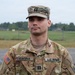 A U.S. Army Captain named Farina is photographed in a formal portrait-style image while wearing Operational Camouflage Pattern (OCP) uniform. He is standing outdoors on what appears to be a military installation, with an open field and tree line visible in the background. His rank insignia (Captain's double bars) is visible on both his patrol cap and chest.