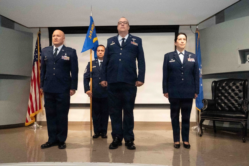 A formal U.S. Air Force military ceremony featuring four personnel in dress blue uniforms standing at attention on a stage. A guidon bearer holds a blue unit flag reading '349 MDG' (Medical Group), flanked by officers with rank insignia and service ribbons. The setting includes a U.S. flag and an Air Force flag, suggesting a change of command or activation ceremony.