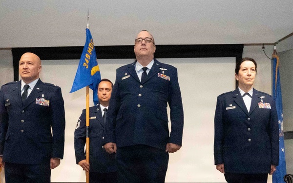 A formal U.S. Air Force military ceremony featuring four personnel in dress blue uniforms standing at attention on a stage. A guidon bearer holds a blue unit flag reading '349 MDG' (Medical Group), flanked by officers with rank insignia and service ribbons. The setting includes a U.S. flag and an Air Force flag, suggesting a change of command or activation ceremony.