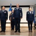 A formal U.S. Air Force military ceremony featuring four personnel in dress blue uniforms standing at attention on a stage. A guidon bearer holds a blue unit flag reading '349 MDG' (Medical Group), flanked by officers with rank insignia and service ribbons. The setting includes a U.S. flag and an Air Force flag, suggesting a change of command or activation ceremony.