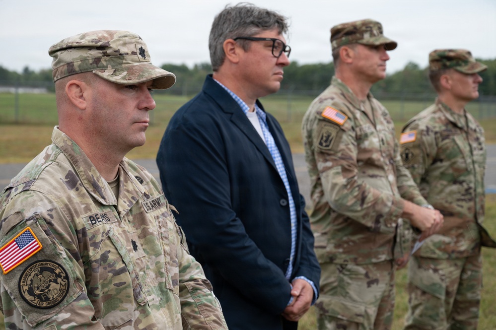 Four individuals stand in a line outdoors at what appears to be a military installation or airfield. Three are wearing U.S. Army OCP (Operational Camouflage Pattern) uniforms, while one civilian in a dark blazer stands among them. The group appears to be observing a ceremony or event.