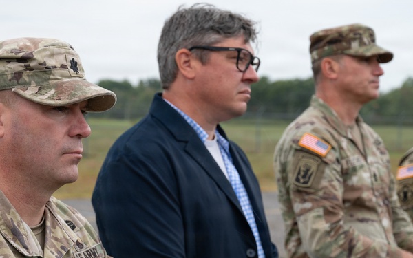 Four individuals stand in a line outdoors at what appears to be a military installation or airfield. Three are wearing U.S. Army OCP (Operational Camouflage Pattern) uniforms, while one civilian in a dark blazer stands among them. The group appears to be observing a ceremony or event.