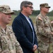 Four individuals stand in a line outdoors at what appears to be a military installation or airfield. Three are wearing U.S. Army OCP (Operational Camouflage Pattern) uniforms, while one civilian in a dark blazer stands among them. The group appears to be observing a ceremony or event.